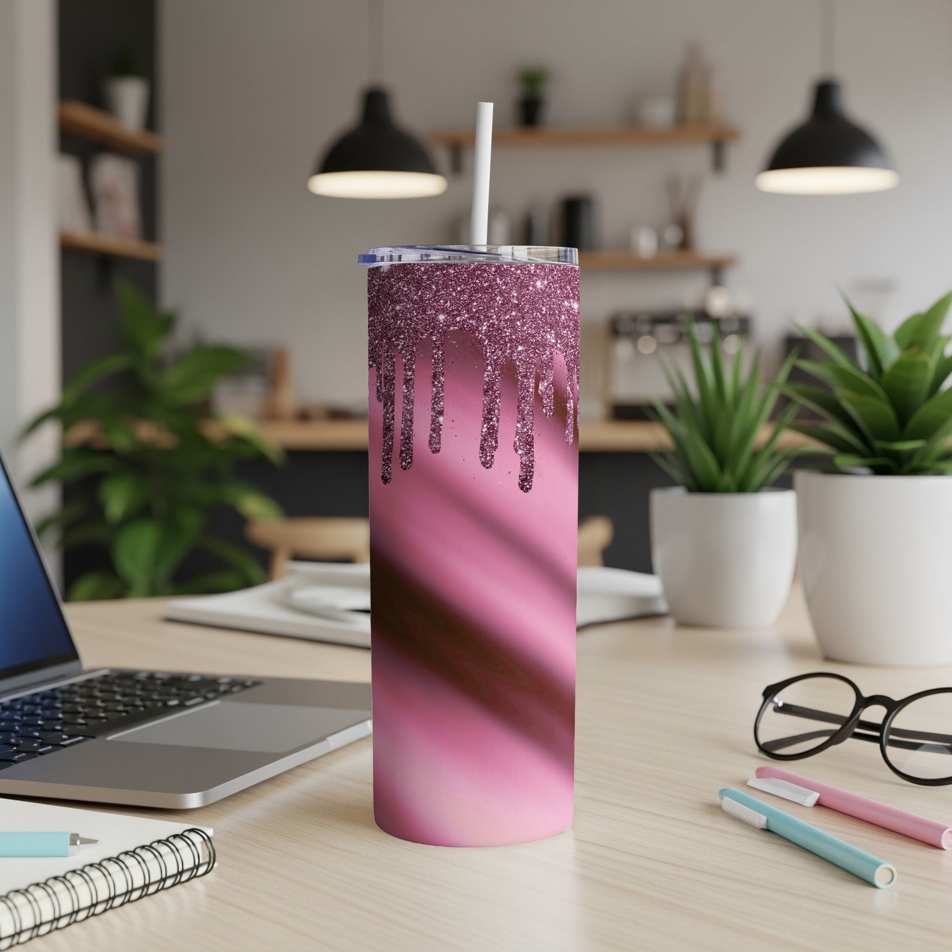 Pink glitter dripping tumbler with straw on a desk beside laptop and glasses.