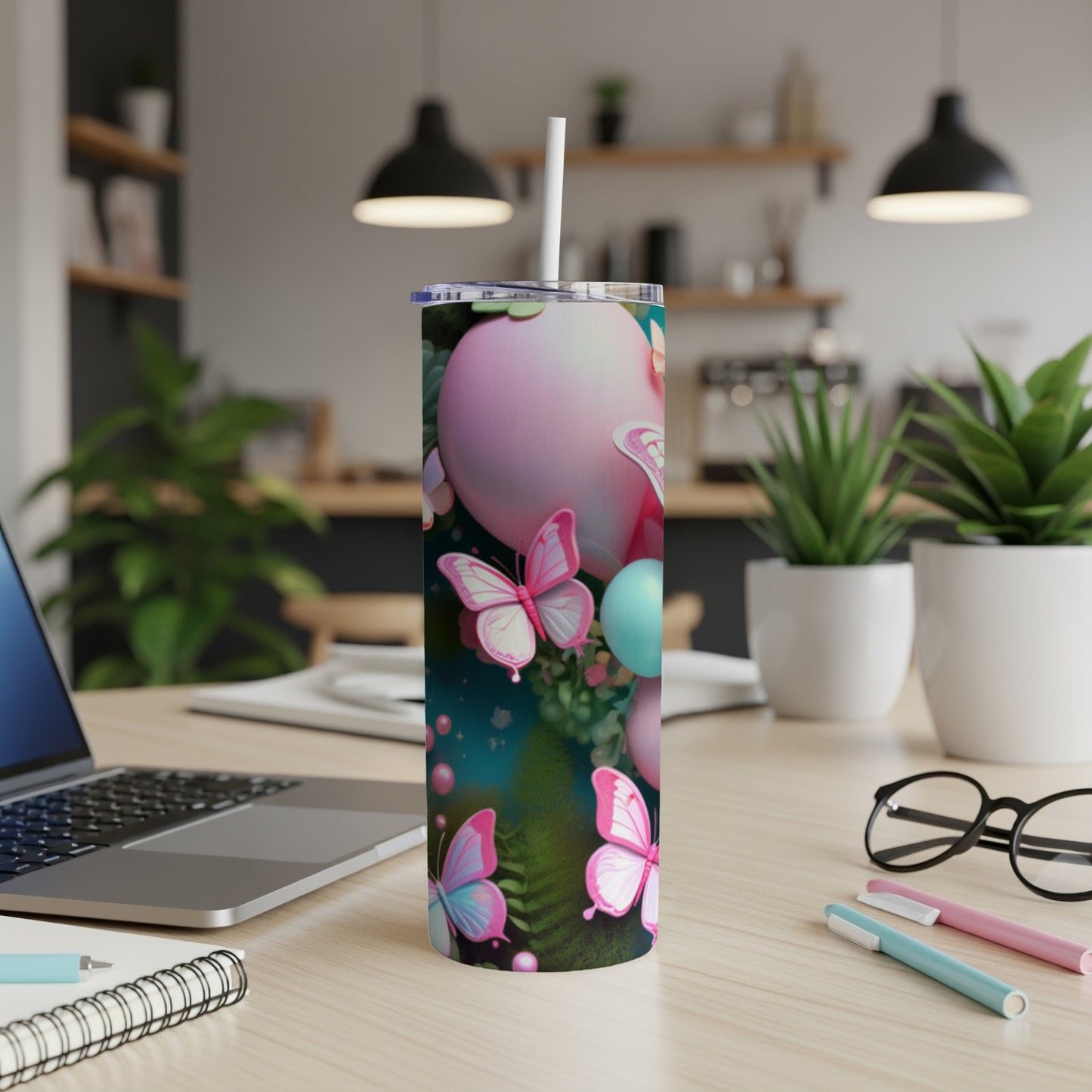 Pink tumbler with butterflies and spheres, next to laptop and pens.