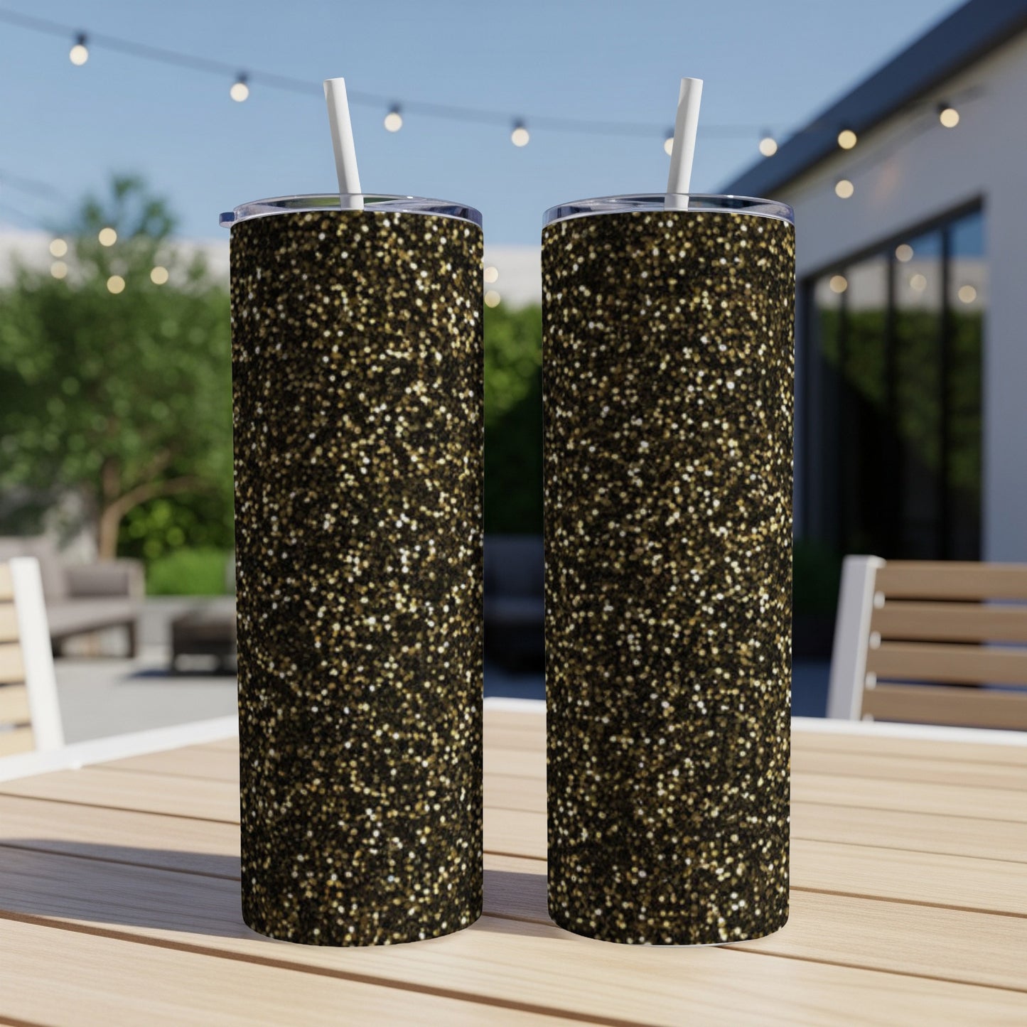 Two tall tumblers with a black and gold glitter pattern sit on a wooden table outdoors.