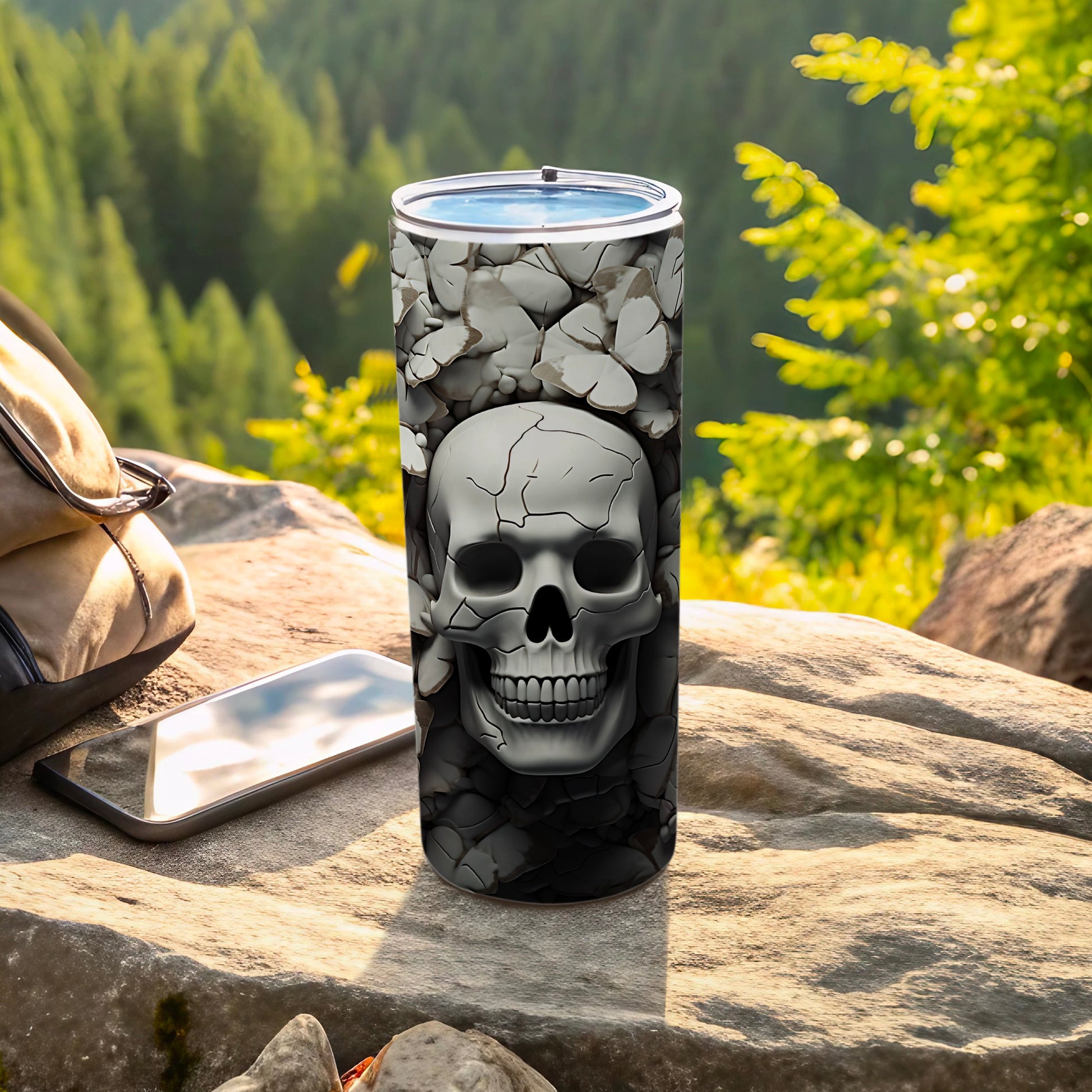 Skulls and rocks design on a tall tumbler with phone and bag outdoors.