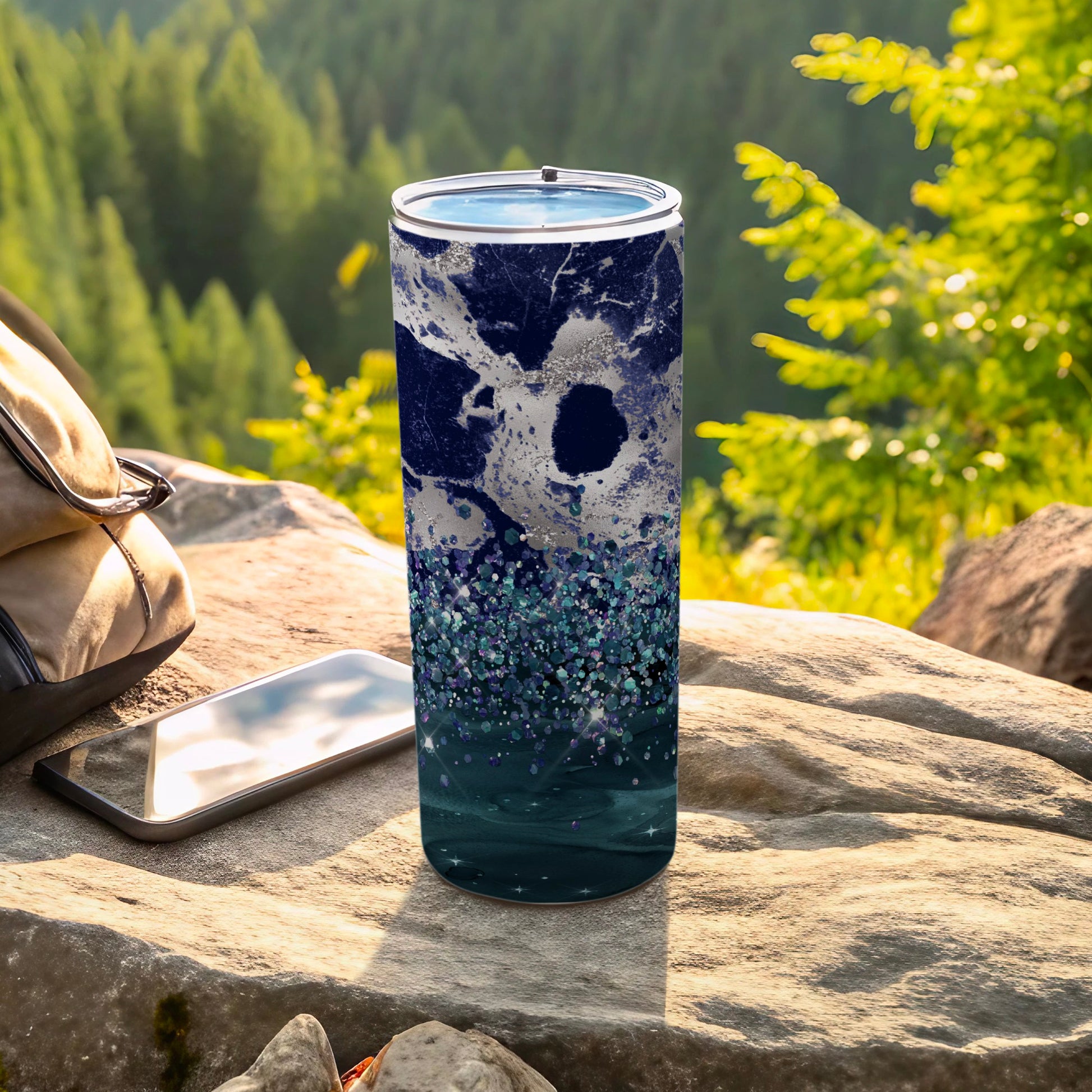 Blue marble and glitter tumbler with lid on rock near phone and backpack.