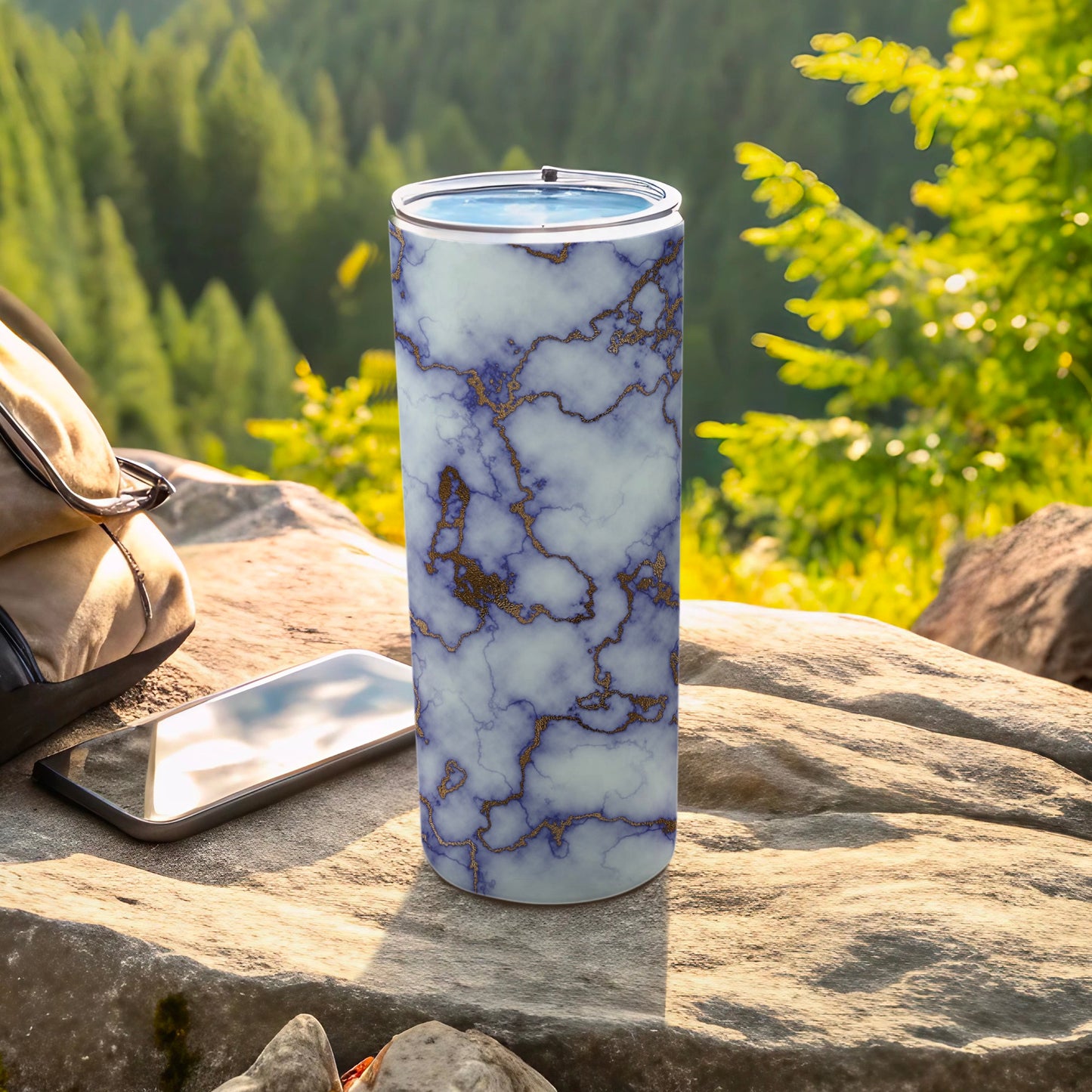 Marble-patterned tumbler with gold accents on a rock with a phone and bag.