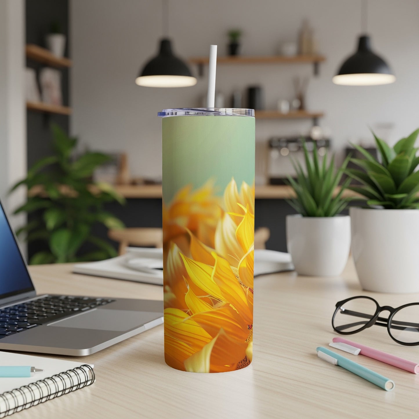 Sunflower-patterned tumbler with straw on a desk next to a laptop and pens.