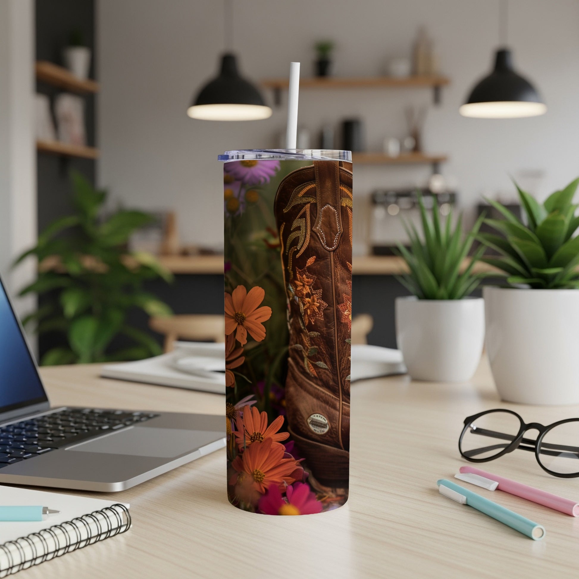 Cowboy boot with floral embroidery on a tumbler, next to a laptop.