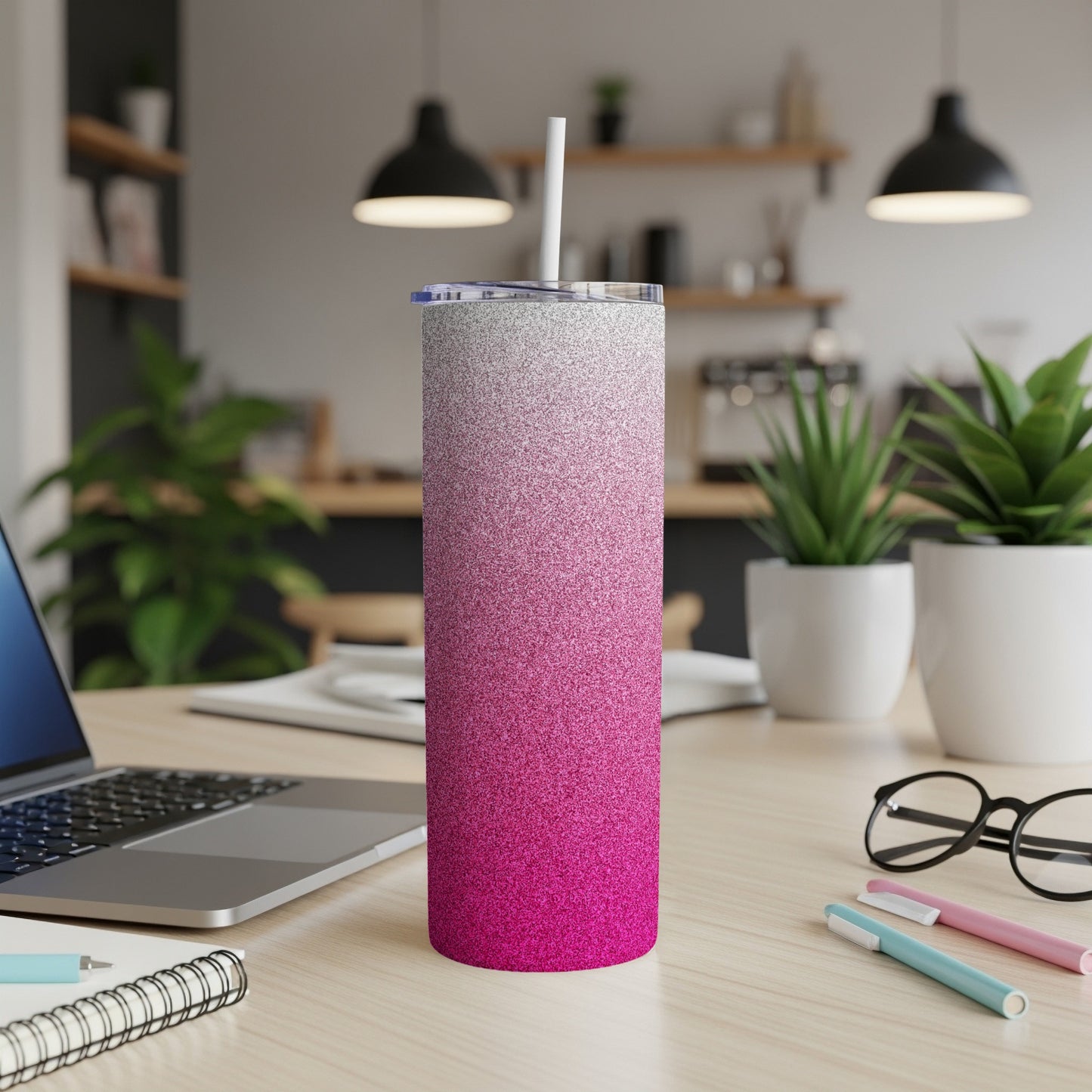 Pink and silver glitter tumbler with straw on desk beside laptop and pens.