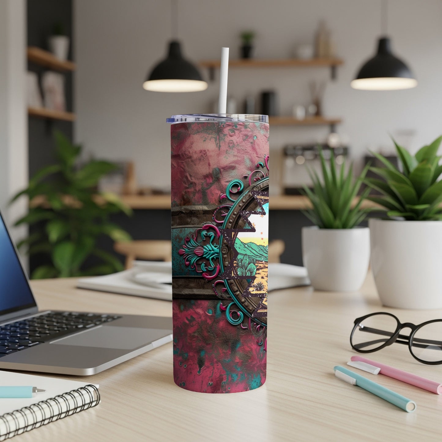 Tumbler with ornate frame and colorful landscape, on desk with laptop and pens.