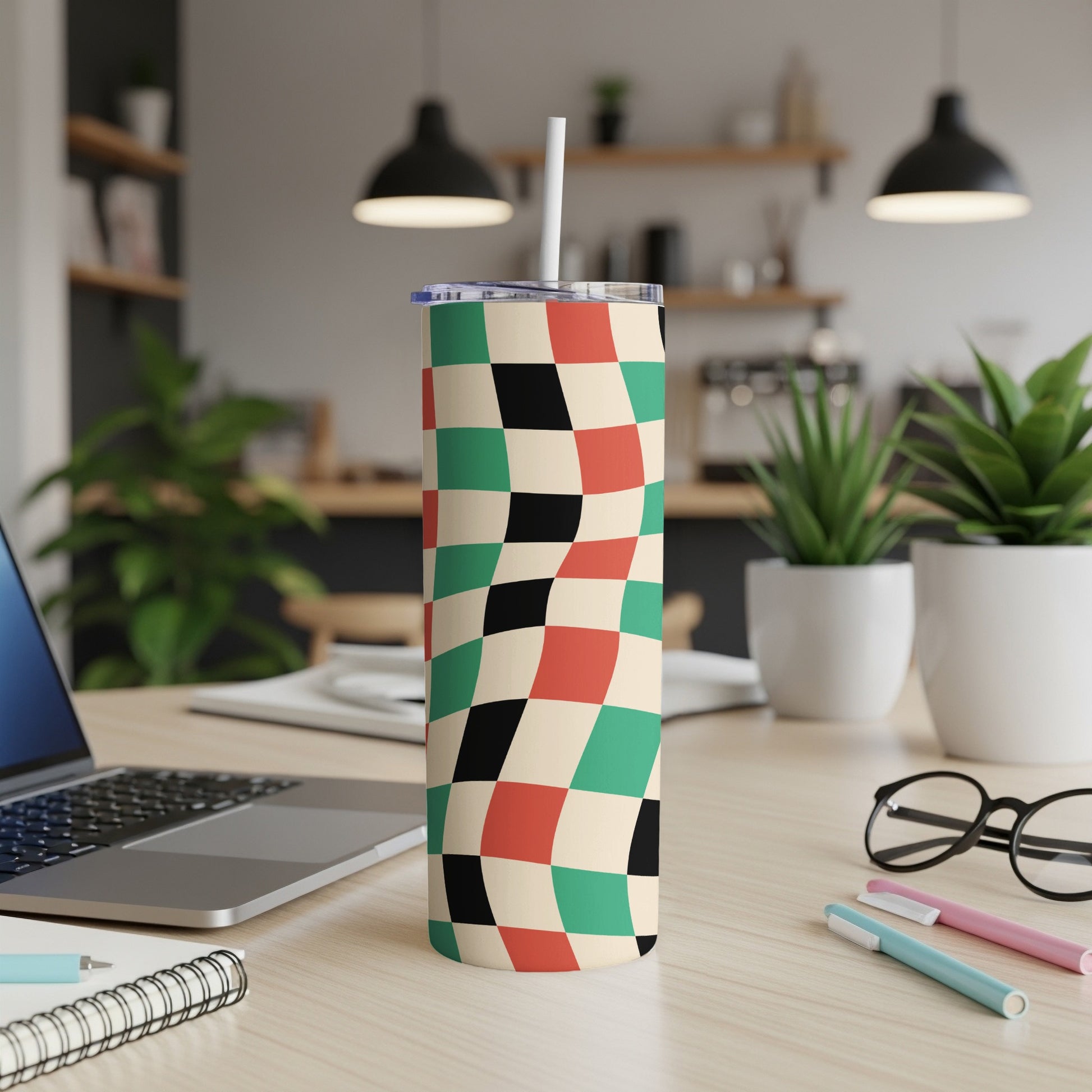 Checkered tumbler with straw on desk next to laptop and pens.