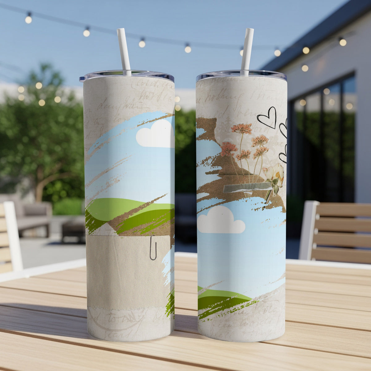 Two tumblers with sky, cloud, and floral designs sit on a wooden table outdoors.