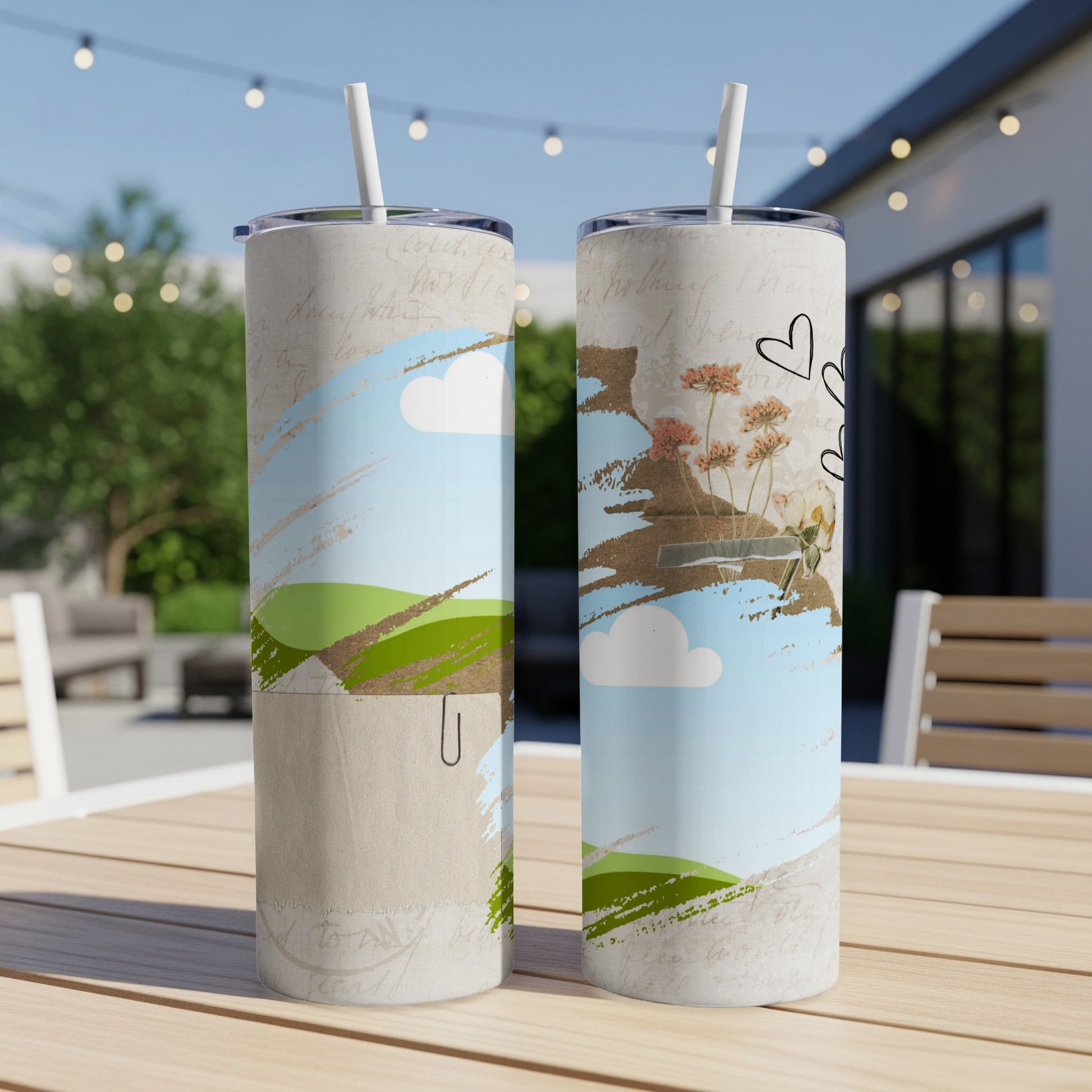 Two tumblers with sky, cloud, and floral designs sit on a wooden table outdoors.