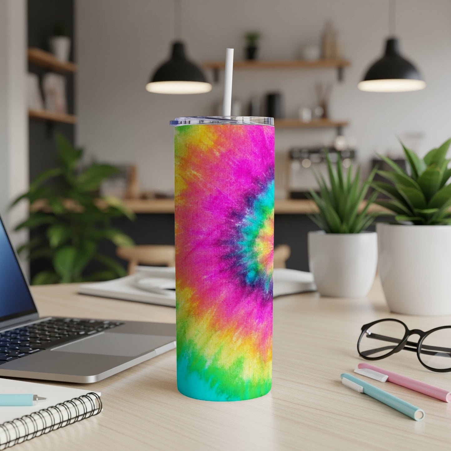 Colorful tie-dye tumbler with straw on desk next to laptop and plants.