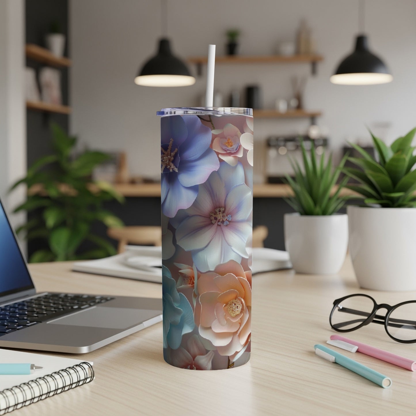 Tall floral tumbler with straw on desk with laptop and pens.