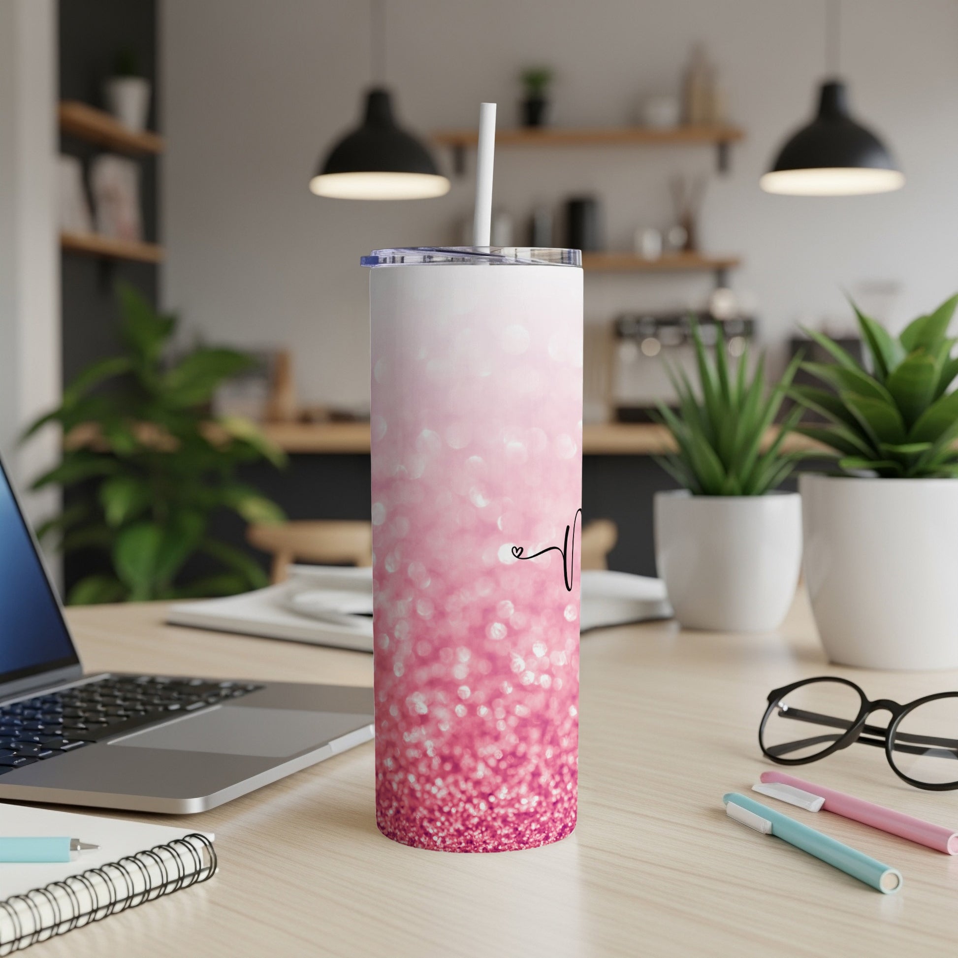 Pink glitter tumbler with a straw and abstract heart design on a desk.