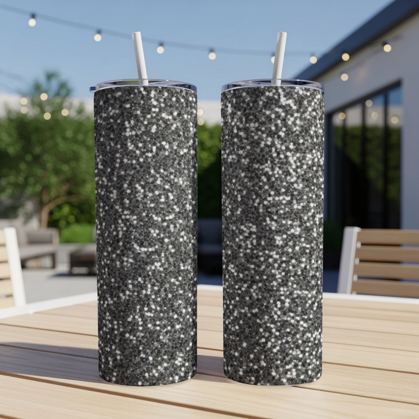 Two glittery black tumblers with straws on a wooden table outdoors.